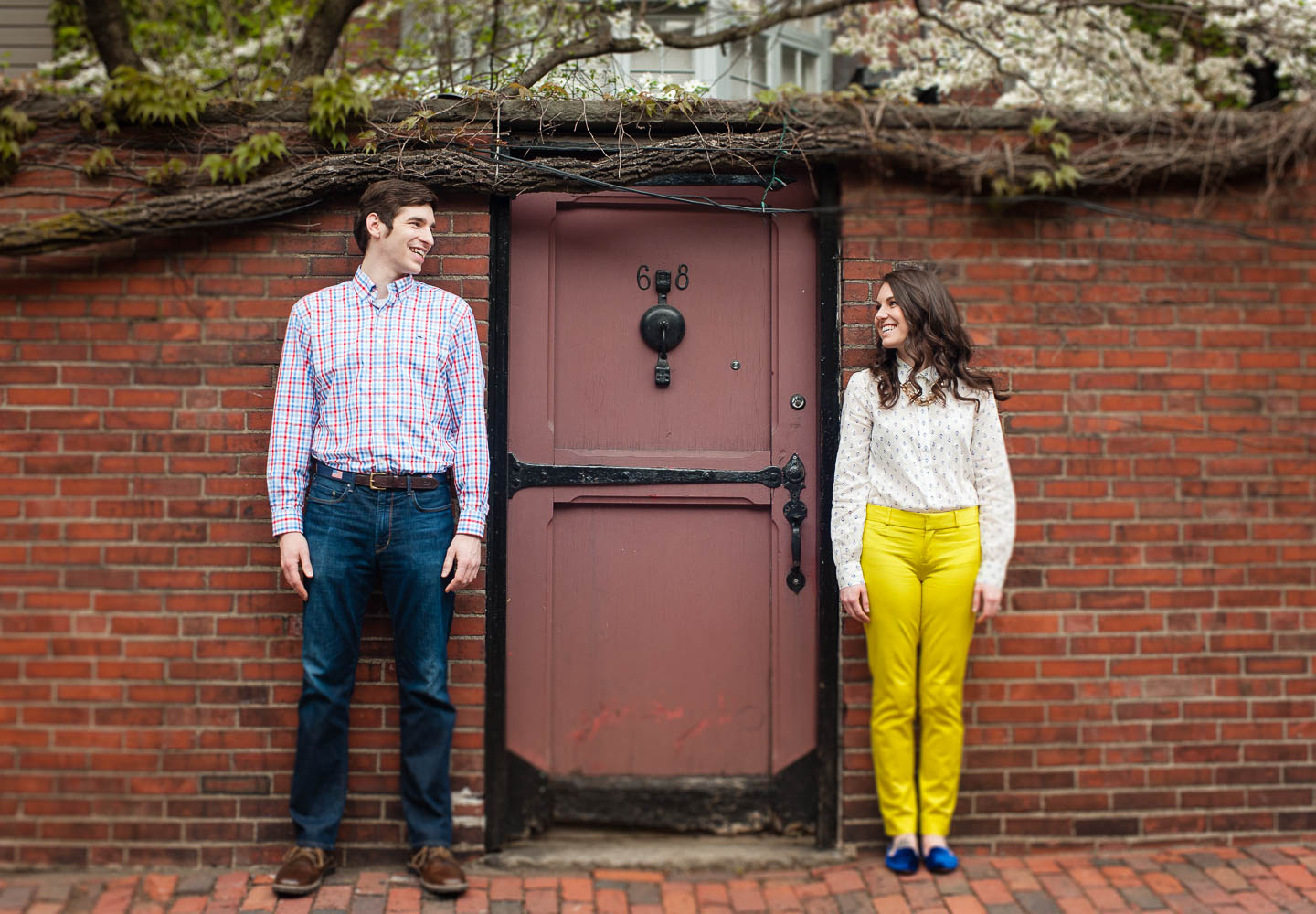 A colorful young couple poses against a deep red brick wall  standing at either side of an iron framed red door during there engagement session