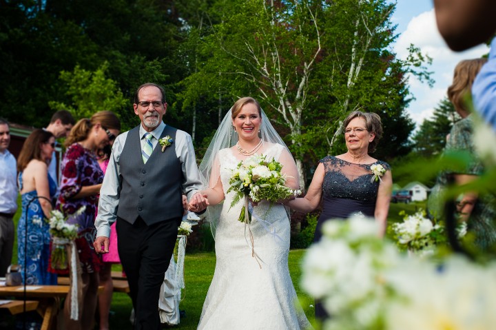 bride and her parents smile at guests while they walk down the aisle of an outdoor ceremony