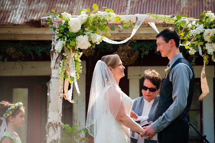 Bride and groom hold hands in from of birch arbor during their barn wedding ceremony