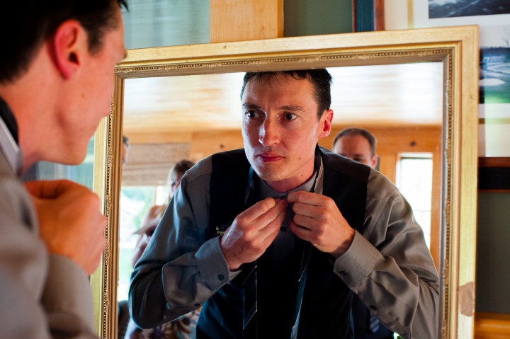 handsome groom buttons collar in the mirror before his rustic barn wedding 