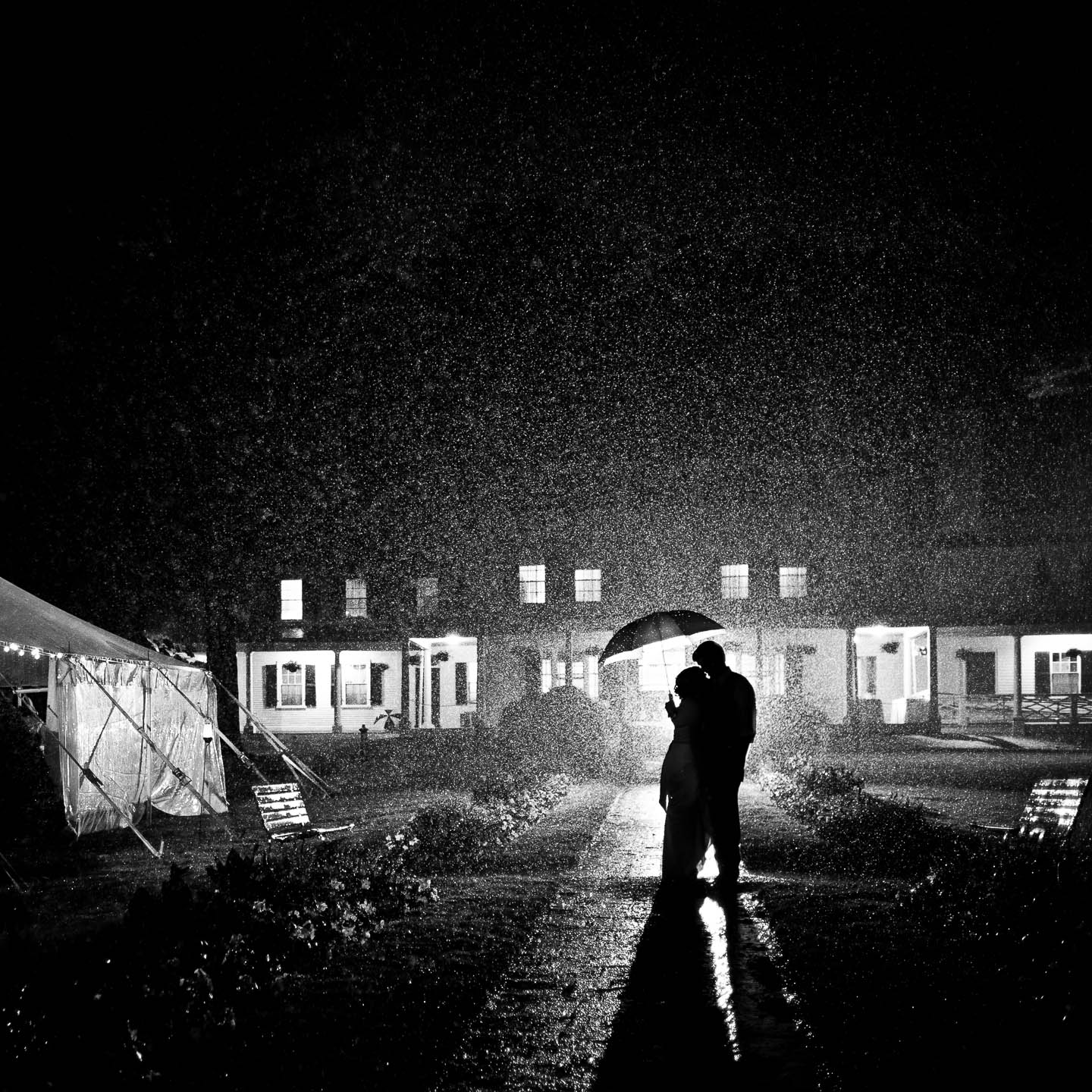 Black and white image of a bride and groom standing under an umbrella in the rain on their wedding day at night in front of their venue