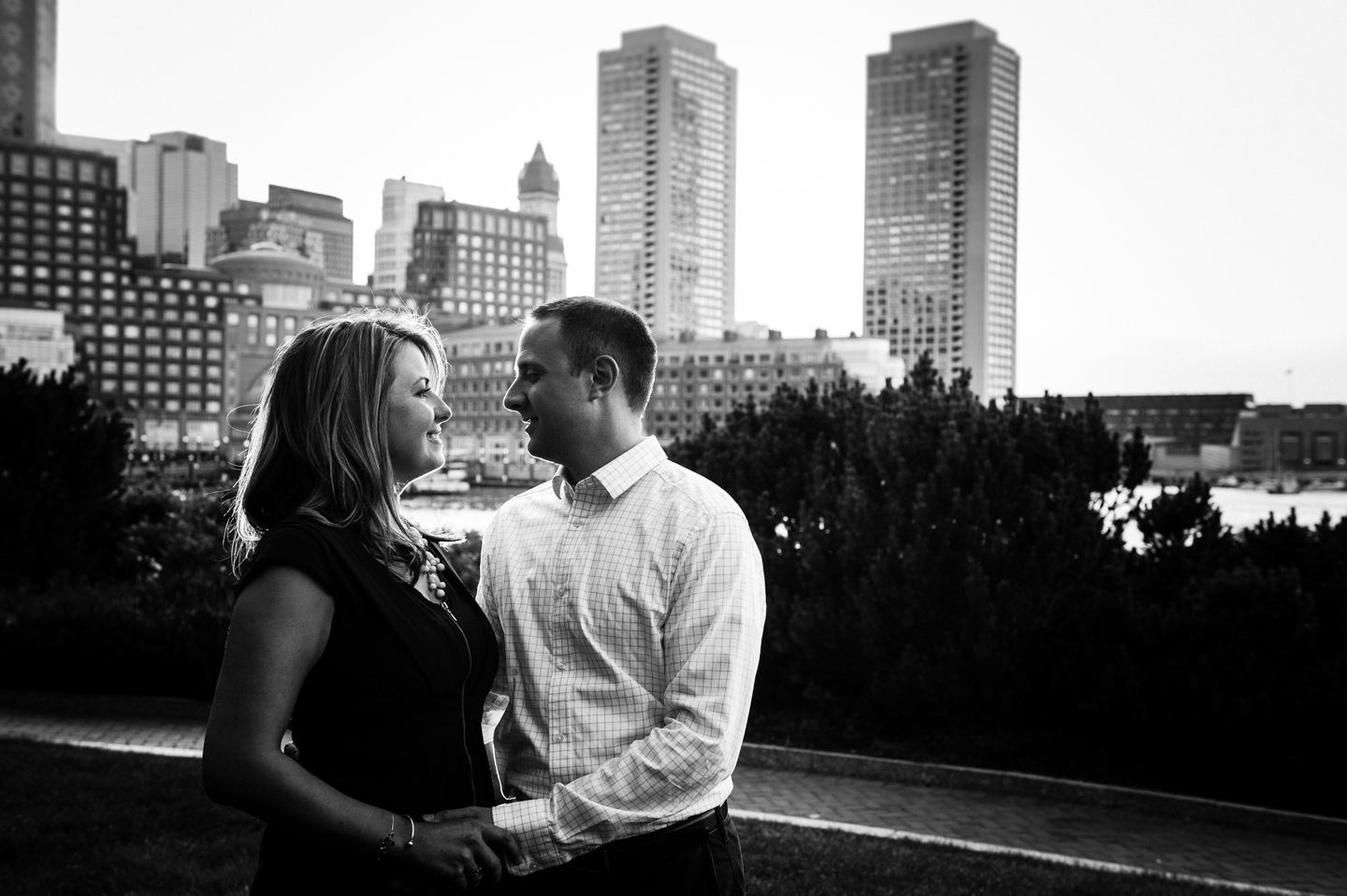 recently engaged couple poses facing each other with the boston city skyline behind 