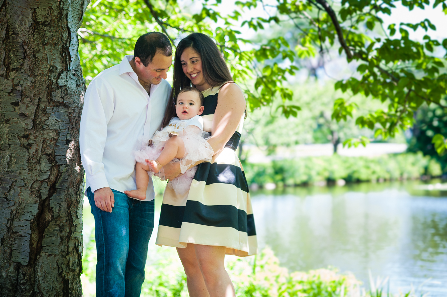 mom and dad pose with their adorable one year old daughter underneath shady trees