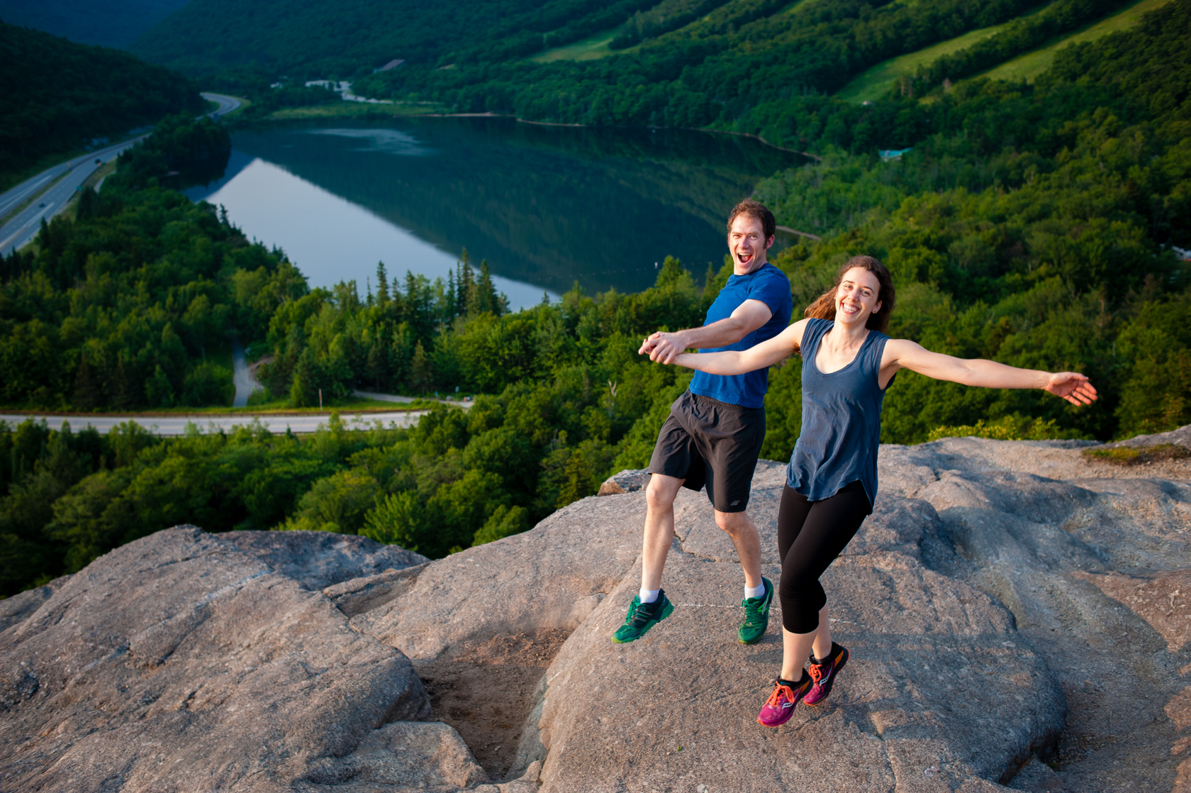 fun couple hold hands and jump during their mountaintop adventure engagement session 