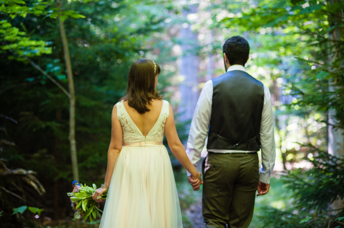 bride and groom hold hands and walk through the forest during their summer mountain wedding 