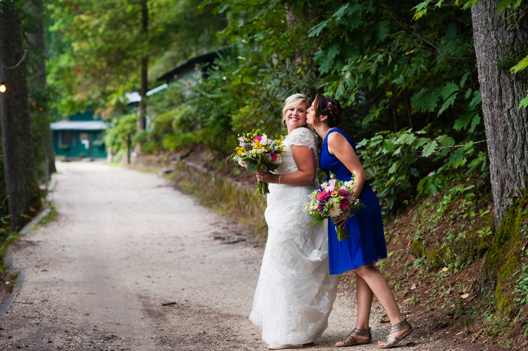 bridesmaid gives beautiful bride a kiss on the cheek on a winding forest lane