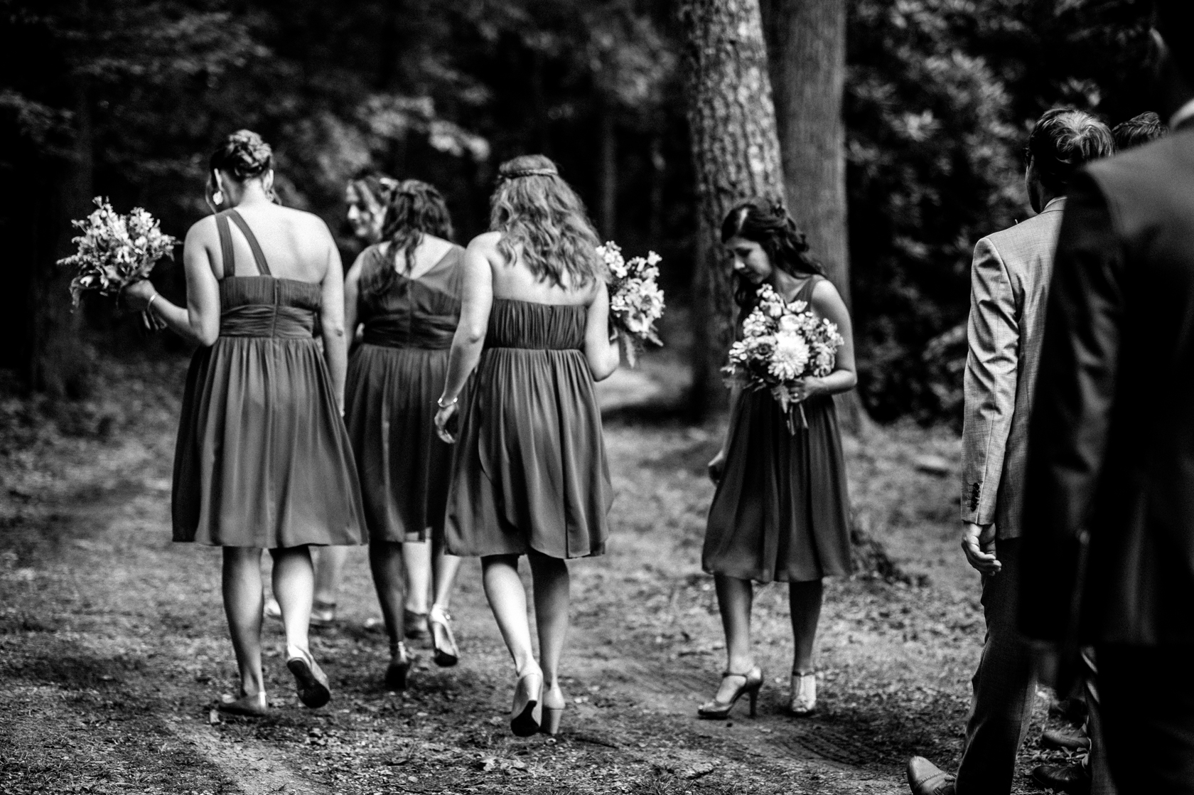 the beautiful bridesmaids wander down a forest path to the ceremony of a summer camp wedding 