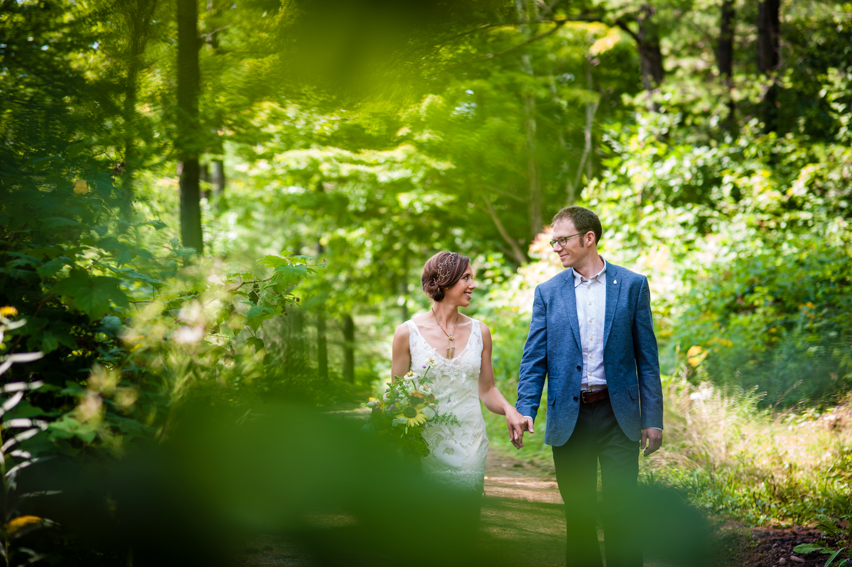 beautiful bride and groom hold hands and walk down a wooded path during their summer camp wedding