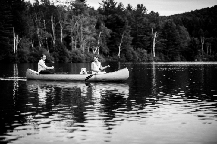 newly married couple paddles a canoe across the lake during their summer camp wedding 