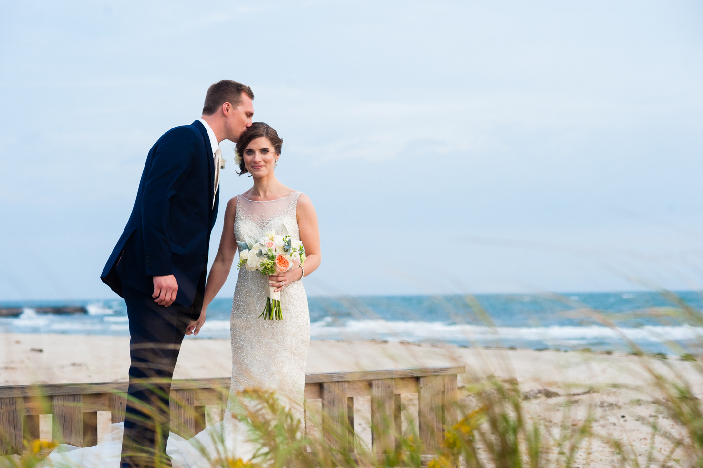 groom gives his gorgeous bride a kiss on the head in the dune grass by the beach 