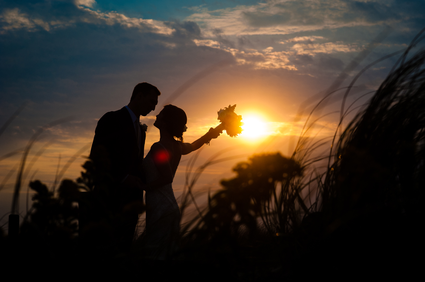 bride raises her bouquet with a gorgeous fall beach sunset silhouetting them 