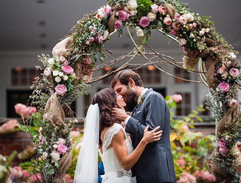 Bride and groom kiss during their wedding ceremony in front of a flower alter